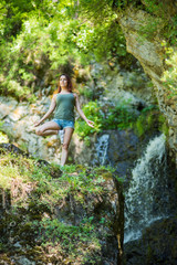 Red-haired woman in shorts doing yoga at the waterfall. A girl stands in a pose of a tree in the forest