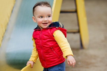 One-year-old boy plays on a playground next to a slide. He is smiling.