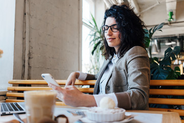 Portrait of handsome curly business woman sitting and working in city cafe with laptop, coffee and cake on the table. Holding mobile phone and texting