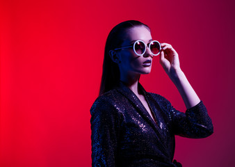Fashion girl with long hair and round sunglasses in a black shining dress poses in neon light in the studio