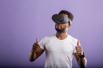 Handsome young African man in VR headset standing against purple background