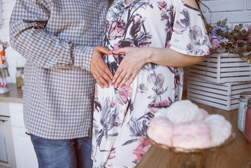 a pregnant girl with her husband, put palms on her belly and formed a heart, the couple is standing in the kitchen with sweets on table and flowers in a vase