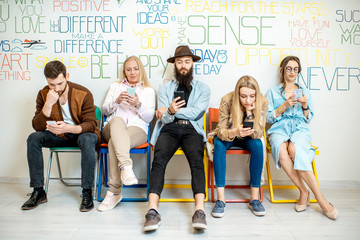 Group of diverse people using smart phones while sitting together in a row on the wall background indoors