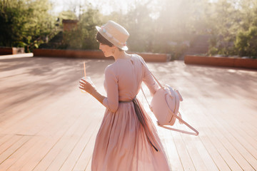 Graceful shapely girl in retro dress dancing outdoor under sunlight, holding trendy backpack....