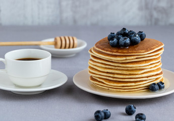 Pancakes with blueberries and honey of acacia on a light plate on a neutral background next to a cup of coffee. In the background is a wooden spoon for honey.