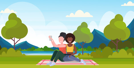 two women taking selfie photo on smartphone camera mix race female characters sitting outdoor on grass posing over nature landscape mountains background flat full length horizontal vector illustration