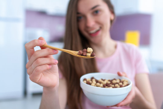 Young, smiling happy beautiful brunette woman holding cereals spoon and cereals bowl full of chocolate balls for breakfast at home