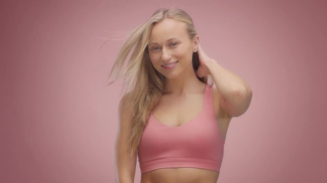 blond model in studio on pink background with hair blowing in air. Touches her neck and hair, smiling and shaking head shaking hair