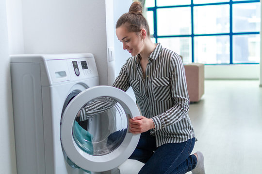 Happy attractive young woman loading laundry in the washing machine at home