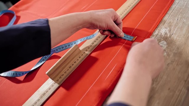 Female's hands are sketching lines with a tape measure and a soap on a table. Sewer works with red fabric and a ruler in atelier. Handicraft concept.