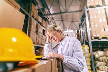 Young blonde Caucasian woman in white uniform and with eyeglasses filling out paperwork while standing in warehouse.