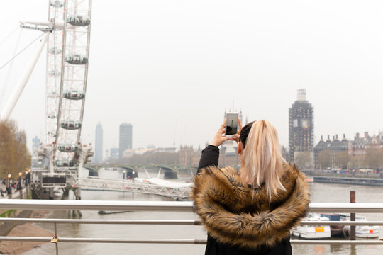 Tourist woman taking picture of London Eye with cell phone by thames river.