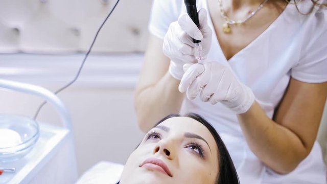 Skilled beautician fiiling up tattoer machine with red ink to make permanent make-up. Cosmetologist preparing for procedure of permanent make-up on lips for a young female in a beauty salon.