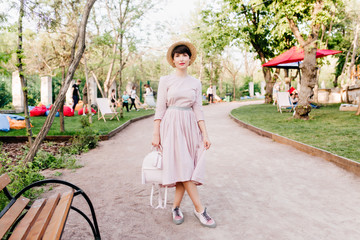 Shapely girl in trendy hat standing with legs crossed near the wooden bench in park. Charming young...