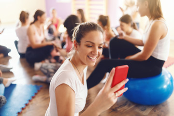 Fototapeta na wymiar Group beautiful fit of sporty women sitting in gym and using smart phones and resting after exercises. Selective focus on brunette with red smart phone.