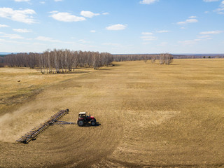 Naklejka premium Aerial of Red tractor with a trailed plow for mowing and weeding fields for the agro-industry of yellow color under the blue sky, a clear spring day. Preparation for planting crops rural technology.