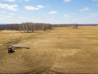 Obraz premium Aerial of Red tractor with a trailed plow for mowing and weeding fields for the agro-industry of yellow color under the blue sky, a clear spring day. Preparation for planting crops rural technology.