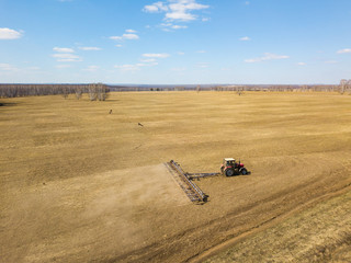 Naklejka premium Aerial of Red tractor with a trailed plow for mowing and weeding fields for the agro-industry of yellow color under the blue sky, a clear spring day. Preparation for planting crops rural technology.