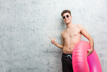 Obraz premium Young man holding a pink inflatable donut wearing a swimsuit excited pointing with forefingers away.
