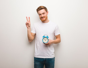 Young redhead man fun and happy doing a gesture of victory. He is holding an alarm clock.