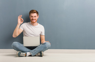 Young redhead student man sitting on the floor cheerful and confident doing ok gesture. He is holding a laptop. - obrazy, fototapety, plakaty