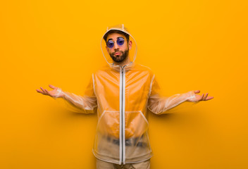 Young man wearing a rain coat doubting and shrugging shoulders