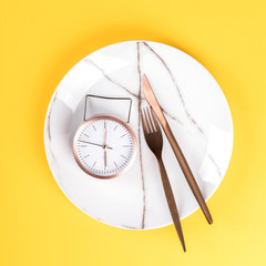 Concept minimal "Time to Eat". Alarm Clock on a white Plate on a on yellow Background.Trendy abstract style.Creative layout. Top View. Flat Lay. Copy space for Text.