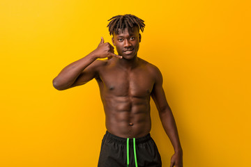 Young black man shirtless wearing swimsuit showing a mobile phone call gesture with fingers.