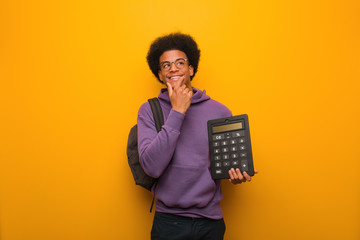 Young african american student man holding a calculator relaxed thinking about something looking at a copy space - obrazy, fototapety, plakaty