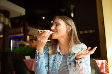 Funny blonde girl in jeans jacket eating pizza at restaurant. 
