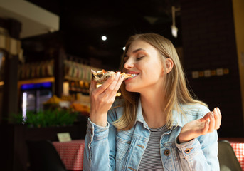 Funny blonde girl in jeans jacket eating pizza at restaurant.  - Powered by Adobe