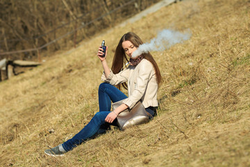 Naklejka na ściany i meble Vape teenager. Young pretty white girl in casual clothing smoking an electronic cigarette on the hillside on a sunny day in the spring. Bad habit. Vaping activity.