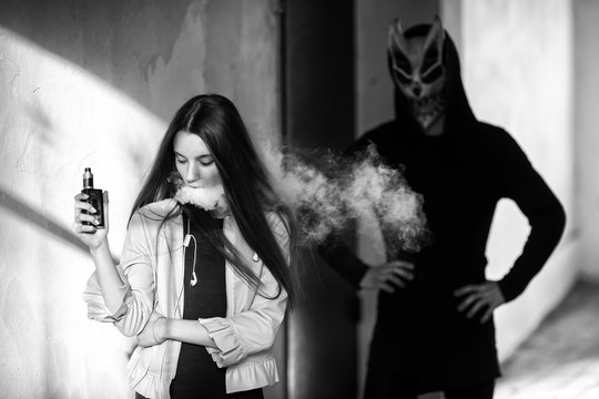 Vape teenager and death. Young cute girl in a dress an electronic cigarette near the wall in front of monster in the background outdoors in spring. Vaping activity. Black and white.