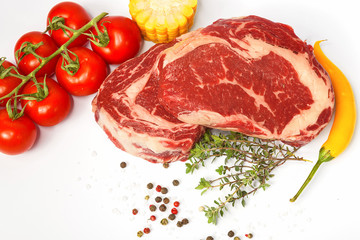 two fresh marbled beef steaks on a white plate with vegetables cherry tomatoes on a sprig, corn, spicy yellow pepper, a sprig of rosemary, salt and different varieties of peppers