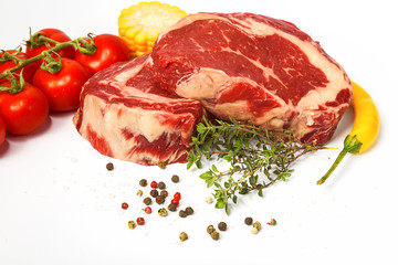 two fresh marbled beef steaks on a white plate with vegetables cherry tomatoes on a sprig, corn, spicy yellow pepper, a sprig of rosemary, salt and different varieties of peppers