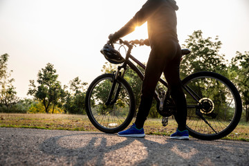 Side view of woman standing with bike in the city park at summer sunset