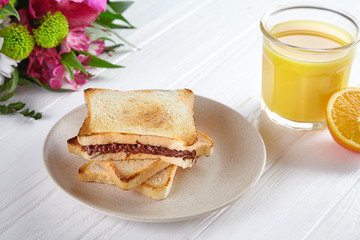 Close up view on toast with Peanut butter. Breakfast concept with orang juice, flowers and sandwich on white wooden background. Traditional summer breakfast. Composition with copy space for design