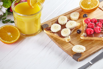Close up view on toast with Peanut butter and raspberries, banana. Breakfast Composition of orang juice, flowers and toast on white wooden background. Traditional summer breakfast. copy space