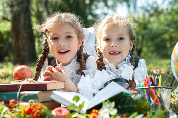 Schoolgirl girls are lying on the grass and reading books. Next to them is a globe, magnifying glass, pencils and an apple.