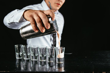 Barman makes white cocktail shots out of shaker. Shooter making shots at a bar. Night out with white alcohol shots on the table. 