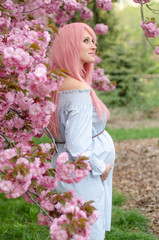 Obraz na płótnie Canvas Pregnant woman in blue dress with pink hair near sakura blooming tree