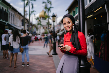 Fototapeta premium A beautiful and young Muslim Middle Eastern woman texts on her smartphone as she leans against a pole on a crowded street with a mosque in the distance. She is smiling confidently.