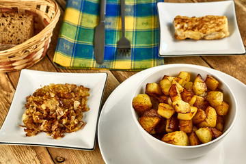Roasted potatoes in a white bowl with rosemary and dill at next to fried onions, fish in batter and cutlery on wooden background. Close-up rustic composition.