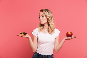 Portrait of hesitating blond woman 20s wearing casual t-shirt holding sweet donut and fresh healthy...