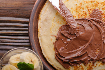 Flat lay French crepes with chocolate sauce and banana in clay bowl on wooden kitchen table