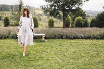  Bohemian woman smiling and enjoying vacation. Atmospheric rustic moment. Copy space. Stylish hipster girl in linen dress and hat walking at lavender field and relaxing in mountains