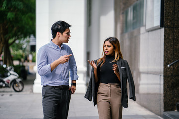 Two diverse Asian businesspeople (colleagues meeting for lunch) walking in the city. One is a Korean man, the other is a Malay woman. They are both seriously speaking as they chat.