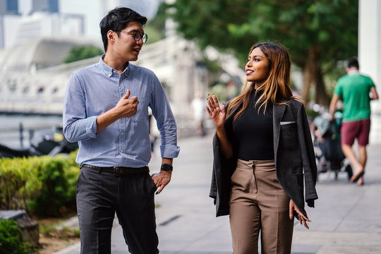 Two Diverse Asian Businesspeople (colleagues Meeting For Lunch) Walking In The City And Is Having An Animated Conversation With Hand Gestures. One Is A Korean Man, The Other Is A Malay Woman.