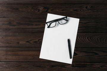 Top view of a white blank sheet of paper with a pen and glasses on a wooden table. White blank paper, pen and glasses on wooden table