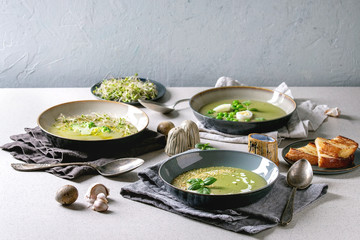 Variety of green vegetable soup asparagus, broccoli and pea, decorated by greens, vegetables, quail eggs, cream, olive oil, in ceramic bowls over grey spotted table with ingredients above.